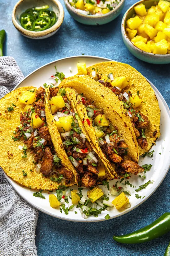 Ein weißer Teller mit zwei Tacos al Pastor, die mit Pineapple, Onion und Tomatillo garniert sind.
