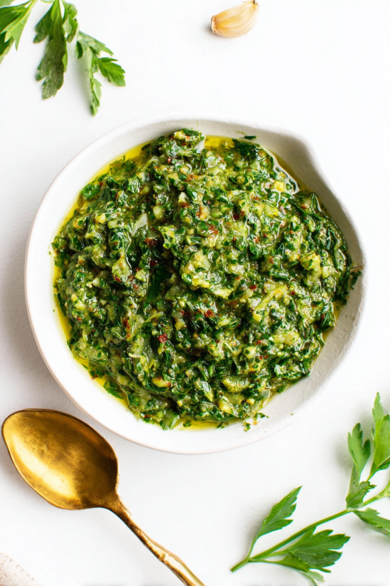 Ein wei&szlig;er Becher mit gr&uuml;nem Chimichurri Sauce, der in einem goldenen L&ouml;ffel gereicht wird.
