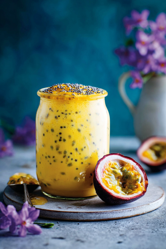 Ein Glas mit einer cremigen Masse, die aus Passionsfrucht und Gewürzen besteht. Es ist auf einem Tisch, der auch eine Schüssel mit Früchten und eine Glasscheibe enthält.