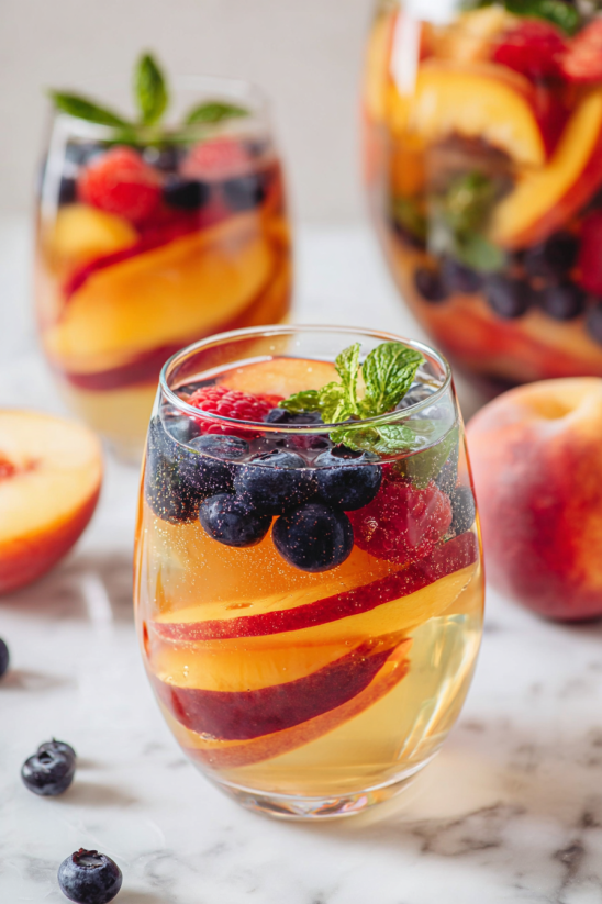 Ein Glas mit frischem Obst, darunter Raspberries, Blaubeeren und Peach, wird von einer Mintgarnitur umgeben.